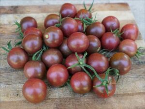 A collection of dark red tomatoes with green stems, matching the rich colors of the Tomato 'Black Cherry' 4" Pot, lies on a wooden surface.