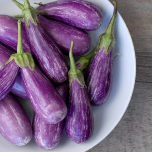 A white bowl filled with 'Fairy Tale' purple eggplants, each with green tops, sits on a wooden surface, echoing the charm of an enchanted pot.