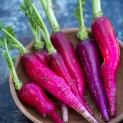 A wooden bowl holds six fresh radishes with green tops, resembling a vibrant 'Fire Candle' on a textured dark surface.