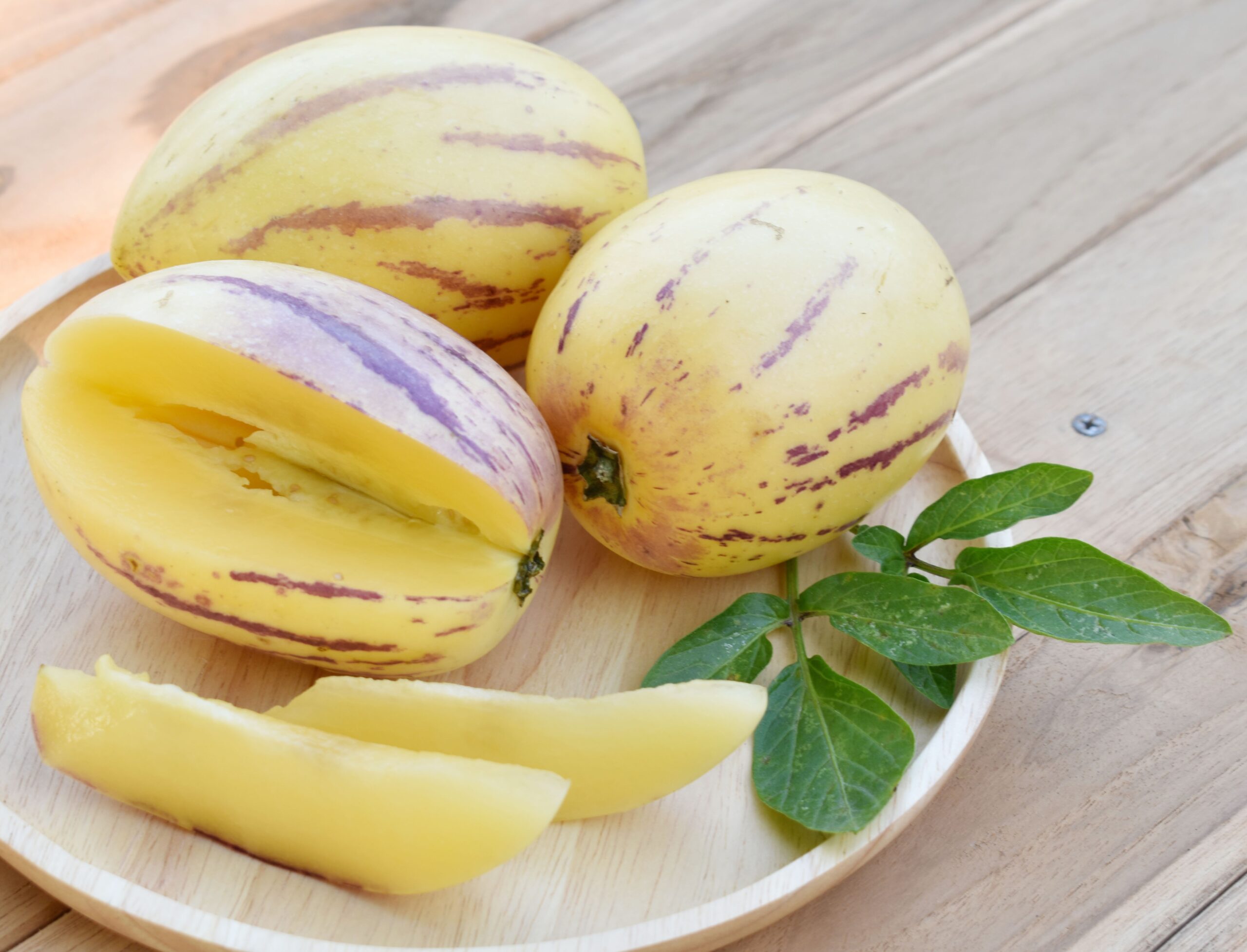 Three Solanum 'Pepino Gold' melons, one sliced open, are displayed on a round wooden plate with a green leaf, placed on a wooden surface. Available as Solanum 'Pepino Gold' 4" Pot.