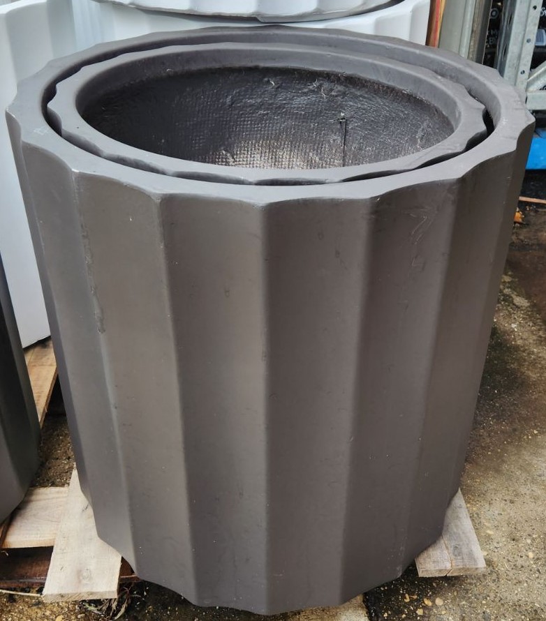 The GardenLite Grecian Cylinder Black S 39x39cm planter, featuring a dark matt finish and vertical ridges, is shown outdoors on a wooden pallet.