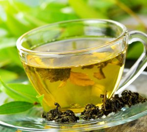 A clear glass teacup filled with green tea, surrounded by loose tea leaves and green foliage in the background.