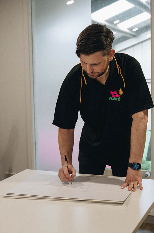 A person wearing a "Hello Hello Plants" shirt stands at a table, writing or drawing on a sheet of paper with a pen beneath a sign that reads, "Bare Rooted Plants for Sale. We're Hiring, Emlyn, staff, employee