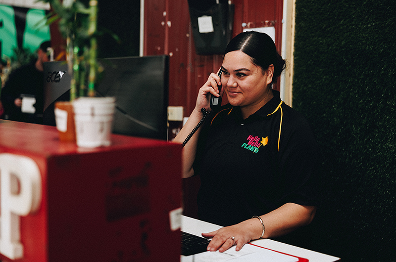 A woman in a black and yellow shirt stands behind a desk, talking on the phone and using a computer. There is a potted plant on the desk with a sign that reads "Bare rooted plants for sale.