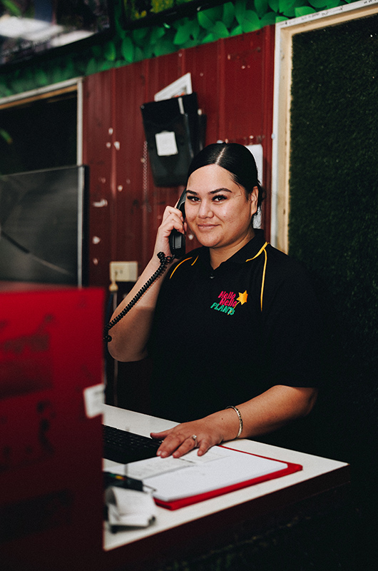 A person, dressed in a black shirt with colorful text and a star emblem, smiles while talking on a corded phone and standing at a counter with a binder, computer, and sign that reads "bare rooted plants for sale.