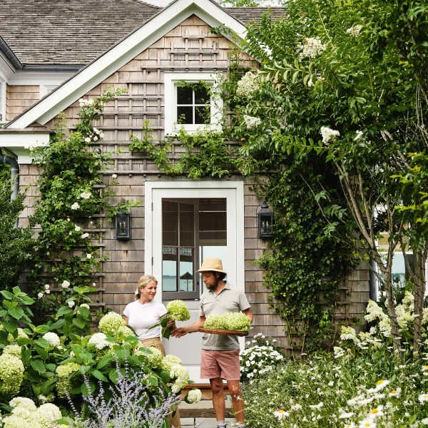 Two people standing in a lush garden, holding hydrangeas, in front of a shingled house with a white door. The garden path is lined with various flowering plants.