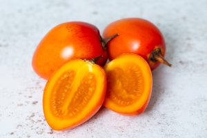 A trio of Solanum 'Denmark Gold' tamarillos, sourced from the Solanum family, are displayed on a textured surface; with two intact and one cut open to showcase its vibrant orange interior.