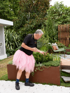 An older man in a black shirt and pink tutu joyfully tends to a raised garden bed in a lush outdoor setting, embracing the many benefits of gardening.