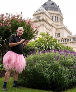 An elderly man in a pink tutu stands by a flowering bush, showcasing the many benefits of gardening, with a historic building gracefully framing the scene.