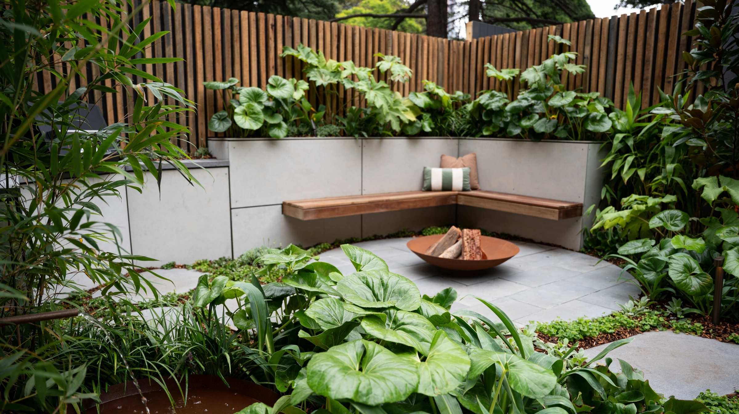 An outdoor garden corner with a wooden bench, enveloped by lush greenery, highlights the many benefits of gardening. A central fire pit warms the circular stone patio, while a vertical wood slat fence completes the backdrop.