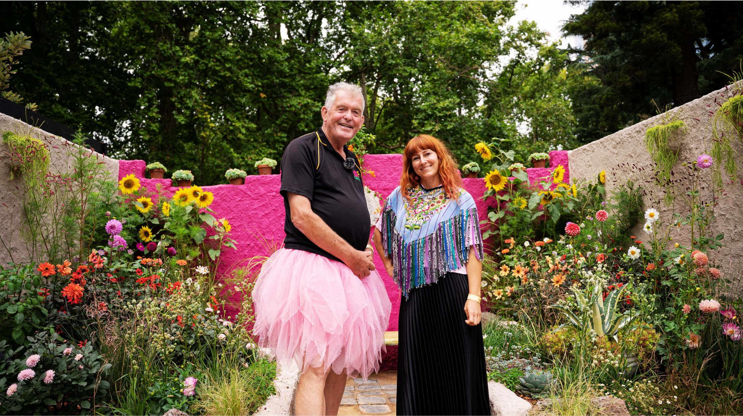 Two people stand in a colorful garden, showcasing the many benefits of gardening. The person on the left wears a pink tutu, while their companion sports a colorful top amidst vibrant foliage and blooming flowers.