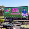 A Hello Hello Plants delivery truck with a green "Hiring" advertisement for free delivery is parked outdoors near garden beds with various plants.