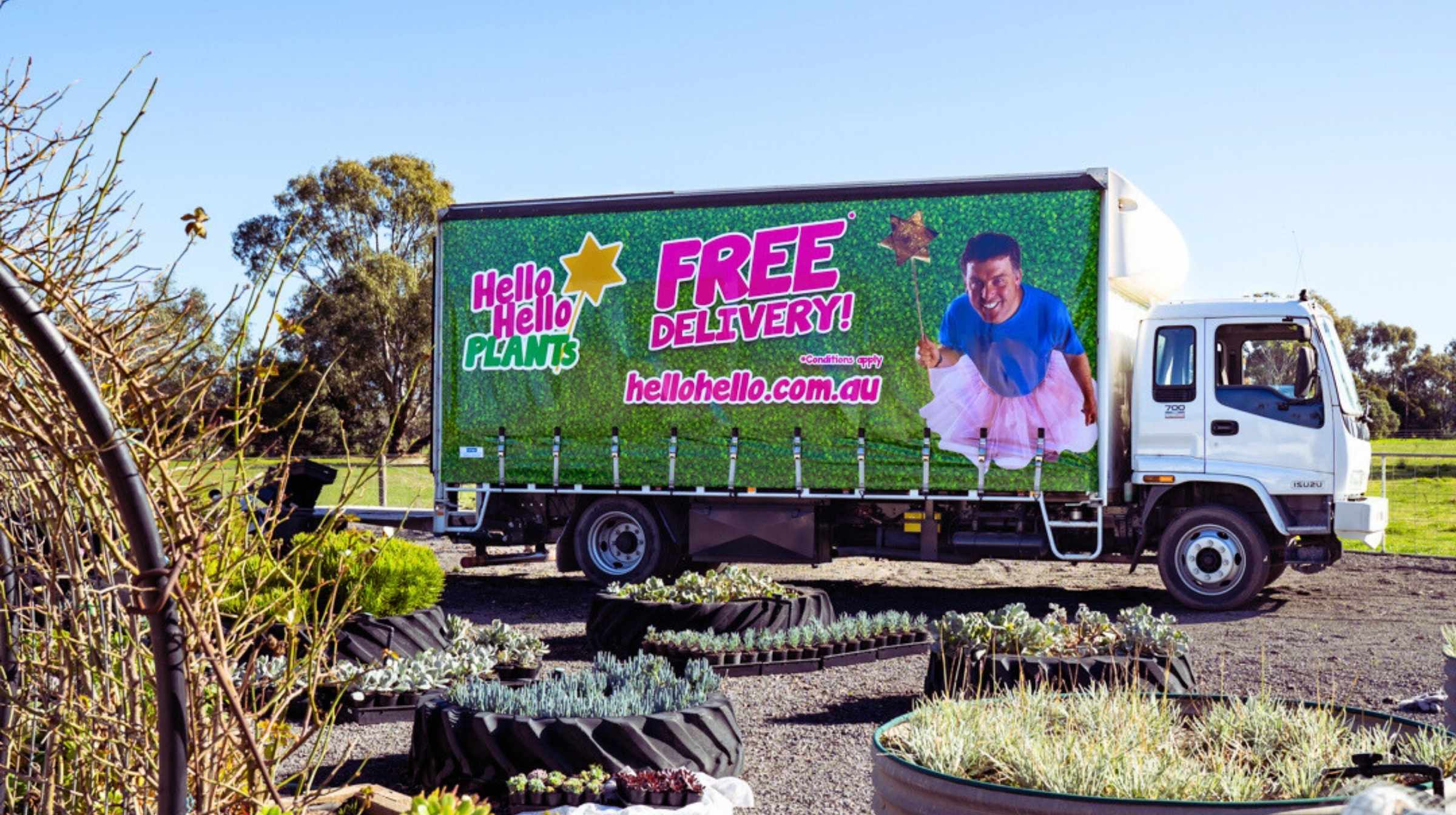 A Hello Hello Plants delivery truck with a green "Hiring" advertisement for free delivery is parked outdoors near garden beds with various plants.