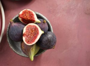 A bowl featuring a mix of whole and halved Ficus 'Violette De Bordeaux' figs sits on a pink surface.