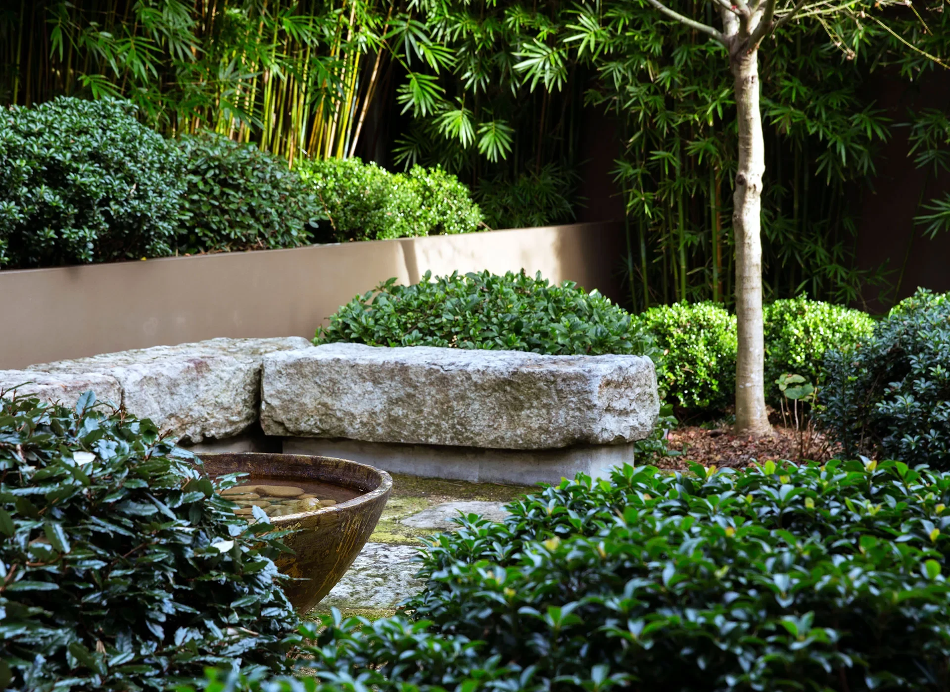 A landscaped garden with dense green shrubs, a tree, a large stone bench, and a shallow bowl water feature in the foreground showcases the beauty of Japanese gardens.
