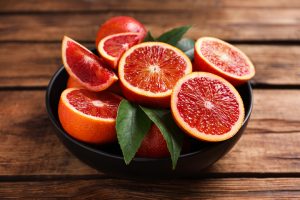 A black bowl filled with blood oranges, some halved and some quartered, with a few green leaves, placed on a wooden surface.