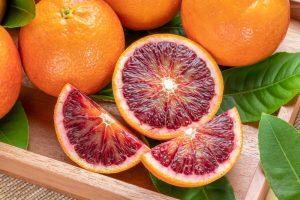 A wooden tray holds whole and sliced Citrus ‘Arnold’ Blood Orange 4L, showing vibrant red flesh, surrounded by fresh green leaves.