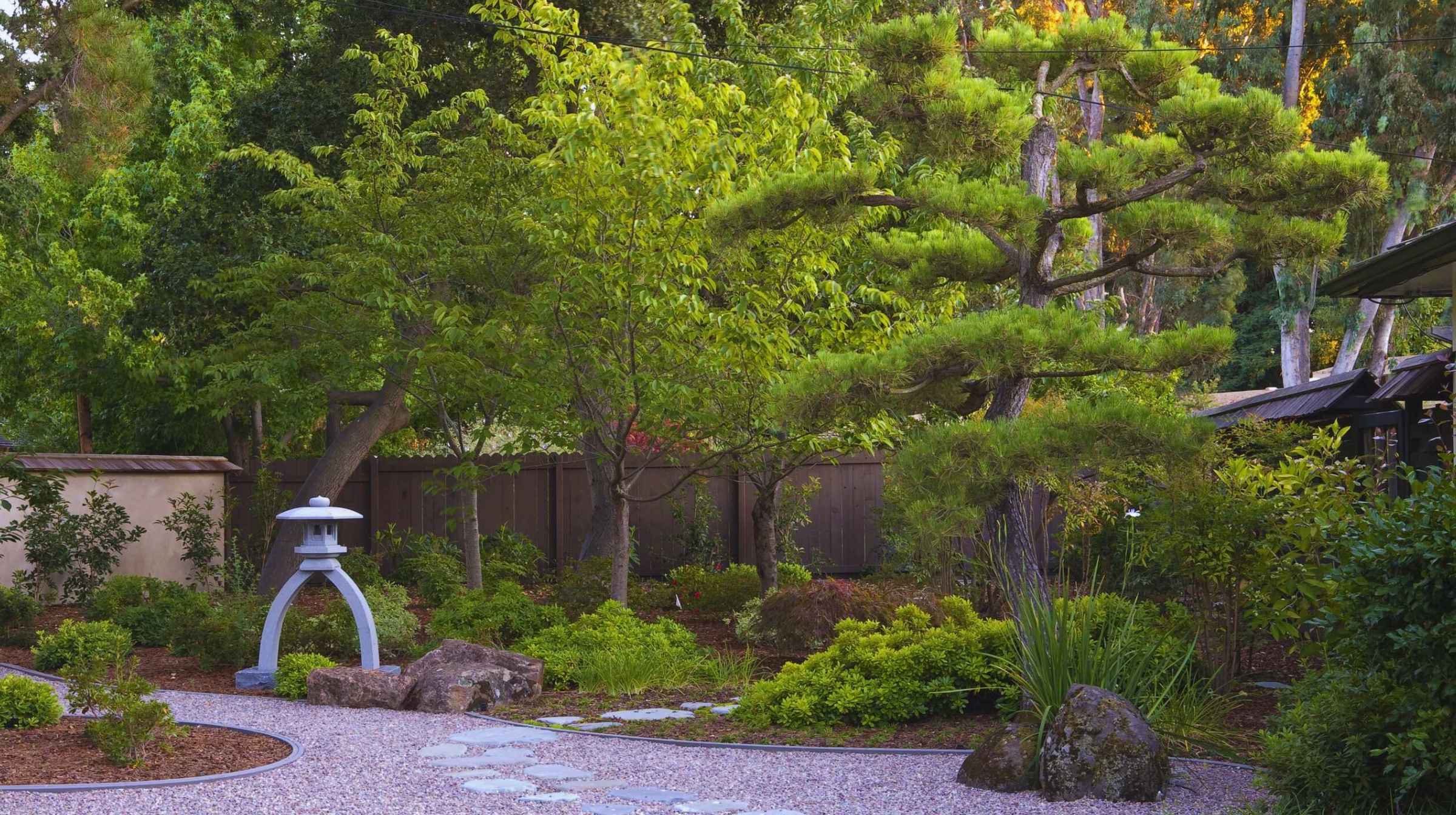 A landscaped Japanese garden showcases the beauty of Japanese gardens with a stone lantern, stepping stone path, manicured trees, shrubs, and a wooden fence in the background.