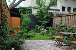 A small backyard garden with a paved patio, wooden table and chairs, green plants, wooden fences, and a tree near a light-colored house captures the tranquil beauty of Japanese gardens in its serene and harmonious design.