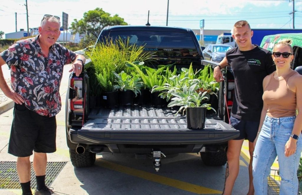 Three people stand by a pickup truck with its tailgate down, displaying various potted plants in the truck bed—ready to help you get the right plants in a convenient parking lot setting.