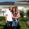 A man, woman, and baby stand together in front of a white house with a garden and hedges on a sunny day, showcasing how curb appeal can increase the value of your property.