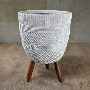 A textured gray concrete planter with geometric patterns stands on three wooden legs against a wooden floor and plain wall background.