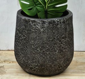 A large, textured dark gray planter with a green monstera plant, placed on a wooden surface against a light-colored wall.