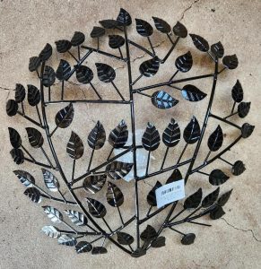 The Raja TeaLight Wall Art Black 46x10cm is a round metal wall decor with a symmetrical tree branch pattern and black leaves, displayed on a concrete surface. A white barcode sticker is near the bottom center.