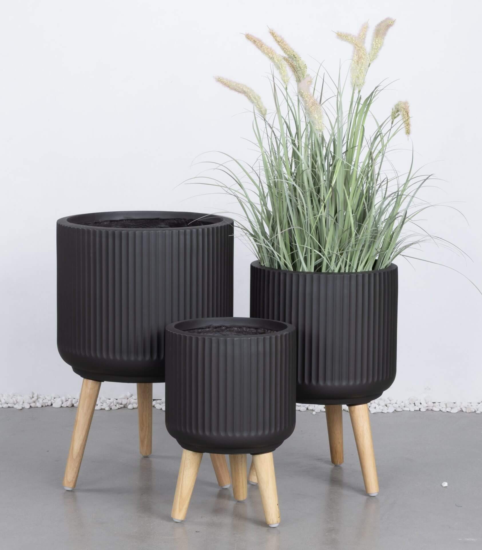 Three black, ribbed plant pots with wooden legs are arranged together; the largest contains tall, green grass-like plants with fluffy tips.