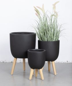 Three black plant pots with wooden legs are displayed on a grey floor; two are empty and one contains ornamental grass with feathery plumes.