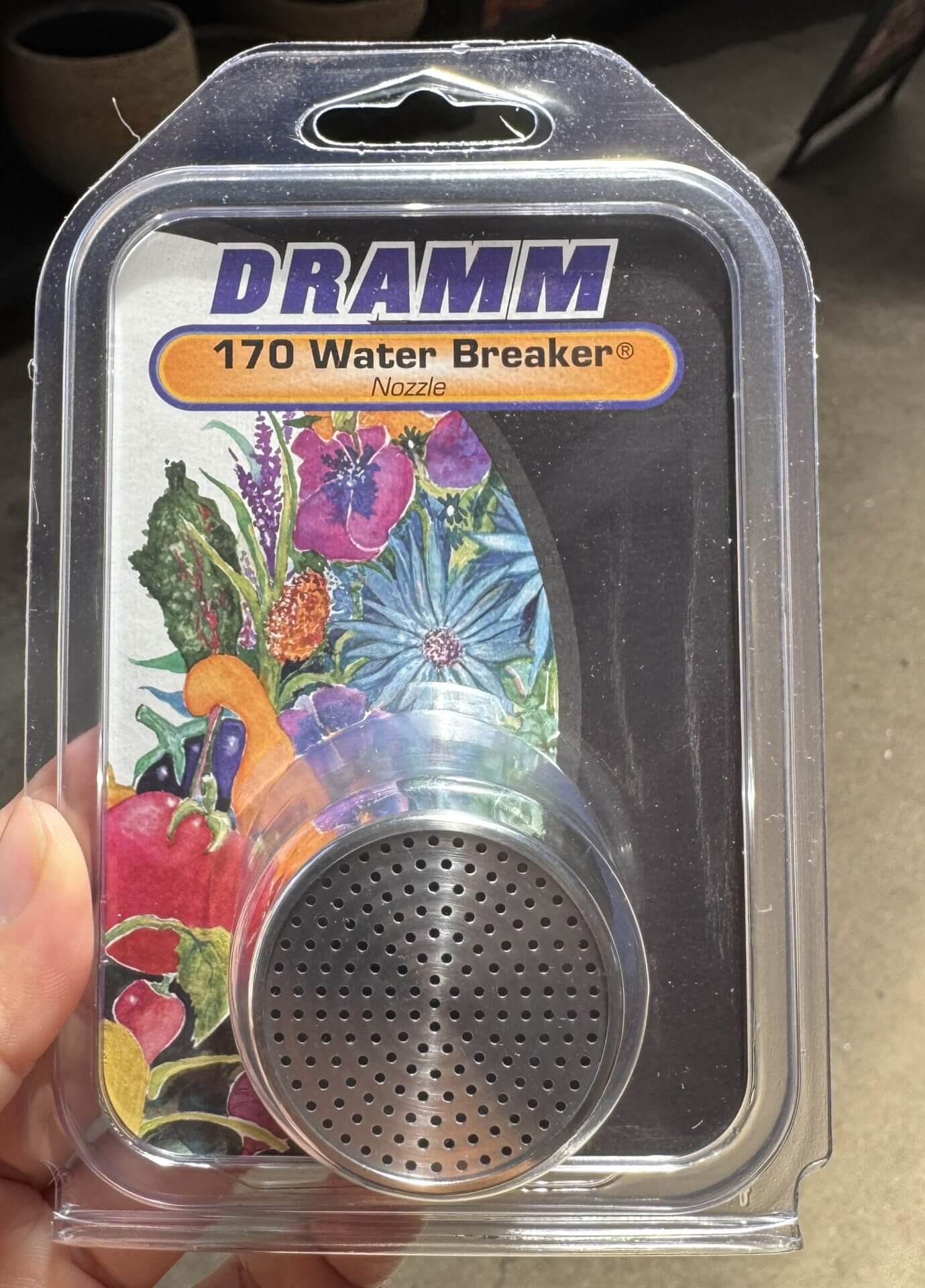 A hand holds a packaged DRAMM 170 Water Breaker Nozzle, featuring a metal spray head and colourful floral artwork on the label.