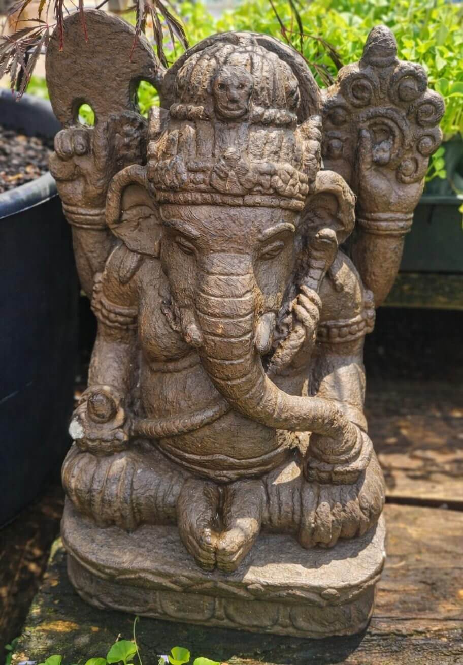 A stone statue of Ganesha, the Hindu deity with an elephant head, seated with one hand raised and detailed carvings, surrounded by greenery.