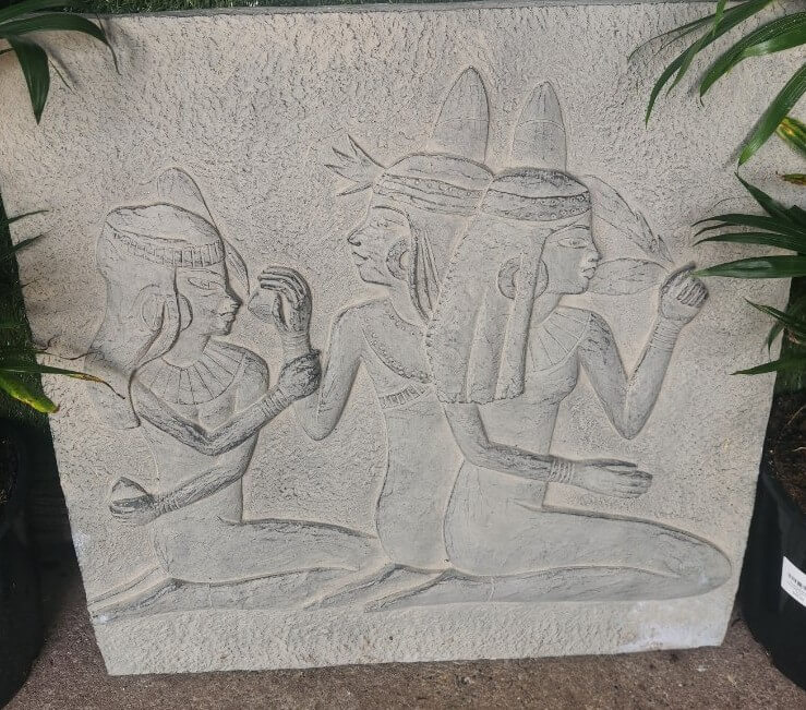 Stone carving depicts three kneeling figures in ancient Egyptian attire and headdresses, with two facing right and one facing left, surrounded by plants on either side.