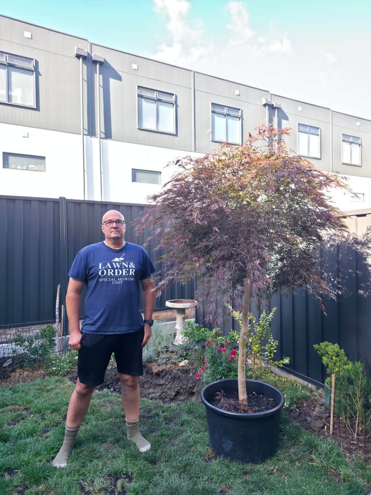 A man in a blue "Lawns & Order" T-shirt stands in a back garden next to a potted tree, with a modern building and garden visible in the background.
