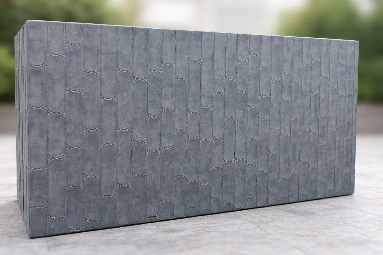 A large rectangular grey concrete wall with a textured, repeating bamboo-like pattern stands outdoors on a paved surface, with blurred greenery in the background.