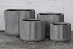 Four grey, ribbed, cylindrical planters of varying sizes arranged on a concrete floor against a white brick wall.