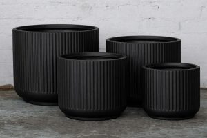 Four black, ribbed cylindrical plant pots of varying sizes are grouped together on a concrete floor against a white brick wall.